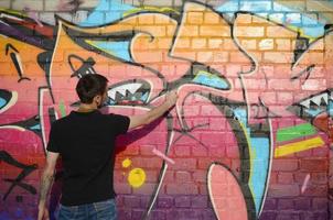 jovem grafiteiro com mochila e máscara de gás no pescoço pinta graffiti colorido em tons de rosa na parede de tijolos. arte de rua e processo de pintura contemporânea foto