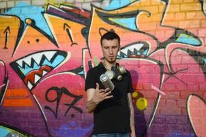 jovem grafiteiro com máscara de gás no pescoço joga sua lata de spray contra graffiti rosa colorido na parede de tijolos. arte de rua e processo de pintura contemporânea foto