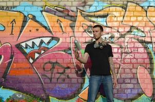 jovem grafiteiro com máscara de gás no pescoço joga sua lata de spray contra graffiti rosa colorido na parede de tijolos. arte de rua e processo de pintura contemporânea foto