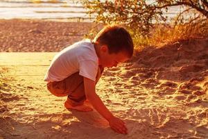 menino brincando com areia na praia. pôr do sol foto