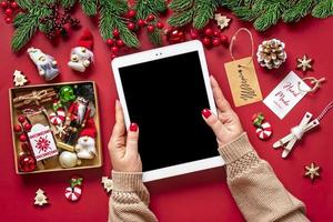 vista superior mãos femininas segurando um tablet com tela digital preta, caixa de natal e decoração na mesa vermelha lista de compras de férias, feliz ano novo, loja online, escolhe presentes, faz maquete de compras foto