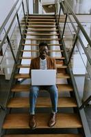 homem de negócios afro-americano jovem trabalhando no computador portátil enquanto está sentado em uma escada de escritório foto