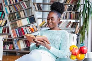 jovem linda garota afro-americana lendo um livro no sofá com as estantes da biblioteca na parte de trás. mulher bonita em um sofá branco lendo um livro foto