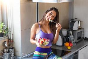 mulher jovem e feliz comendo salada saudável sentada na mesa da cozinha com ingredientes frescos verdes dentro de casa. bela mulher em forma comendo salada saudável após treino de fitness foto