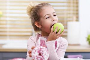 menina bonitinha escolhendo entre maçã e rosquinha na cozinha foto