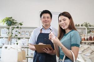 lojista masculino asiático e cliente feminina sorriem e olham para a câmera para produtos naturais em recipientes reutilizáveis na loja de recarga, mantimentos com desperdício zero, varejo orgânico e estilo de vida de compras sustentável. foto