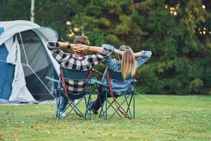 jovem casal simpático sentado em cadeiras na frente da barraca no acampamento foto