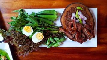 vista superior da comida local tailandesa com carne de porco frita ou grelhada, peixe fermentado, cebola roxa e pimentão verde na tábua de madeira, ovo cozido e vegetais frescos no prato ou prato branco. comida asiática picante foto