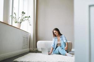 jovem de jeans trabalhando no laptop sentado no tapete em casa, vista das portas foto