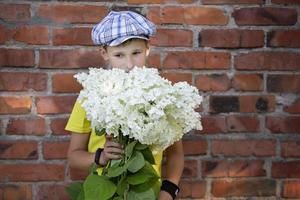 criança alegre e feliz da aldeia com um buquê de flores. menino sorridente com hortênsia perto da parede de tijolos grunge. dia das mães, 8 de março, conceito do dia internacional da mulher. foto