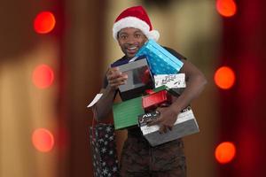 homem usando chapéu de papai noel e segurando muitas caixas de presentes de natal em fundo de luzes vermelhas foto