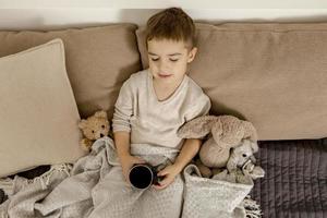 adorável menino bebendo chá quente na cama em casa e relaxando. criança descansando, enrolada em cobertor, com caneca no quarto. interior e roupas em cores naturais da terra. ambiente aconchegante. foto