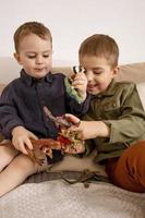 dois garotinhos caucasianos fofos brincando com dinossauros em casa. interior e roupas em cores naturais da terra. ambiente aconchegante. crianças se divertindo com brinquedos, dois amigos. foto