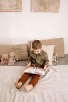 menino caucasiano bonitinho lendo um livro na cama em casa. interior e roupas em cores naturais da terra. ambiente aconchegante. criança lê um conto de fadas. foto
