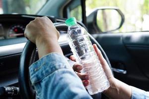 motorista de mulher asiática segura água fria para beber no carro, perigoso e arrisca um acidente. foto