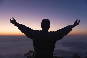 silhueta de um jovem orando a Deus na montanha ao pôr do sol de fundo. levantando as mãos em adoração. conceito de religião cristã. foto