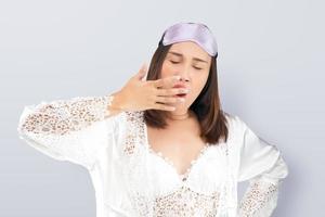 mulher asiática vestindo uma camisola de renda branca e roupão de cetim bocejando mostrando um gesto sonolento cobrindo a boca com a mão. foto