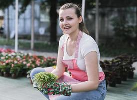 jovem em um viveiro segurando uma planta com flores nas mãos enquanto ela se ajoelha na passarela entre as plantas. foto