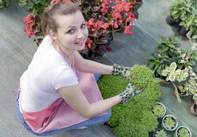 jovem em um viveiro segurando uma planta com flores nas mãos enquanto ela se ajoelha na passarela entre as plantas. foto