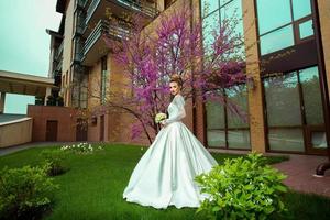 linda jovem noiva loira em vestido de noiva olhando para a câmera ao ar livre na frente da árvore de sakura foto