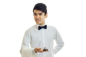 garçom jovem atraente com gravata borboleta e camisa branca segurando um sino e sorrindo foto