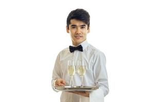 Garçom jovem atraente em uma camisa branca parece sorrisos diretos e segura uma bandeja de copos de vinho foto