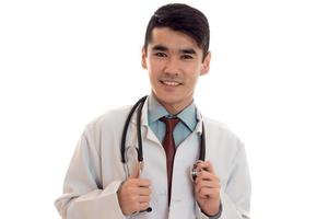 retrato do jovem médico morena feliz de uniforme branco sorrindo para a câmera com o estetoscópio no pescoço isolado no fundo vazio foto