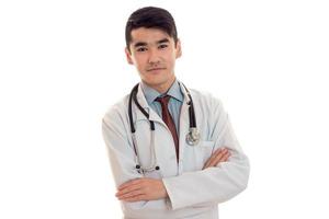 um jovem médico em um jaleco branco com um estetoscópio nos ombros fica com as mãos cruzadas isoladas no fundo branco foto