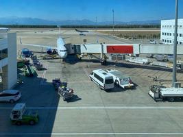um avião com passarela e corredor aguarda partida e carregamento de bagagem no aeroporto e carros de serviço foto