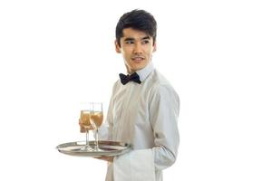 jovem garçom bonito com gravata borboleta e camisa desvia o olhar e mantém as taças de champanhe em uma bandeja que foto