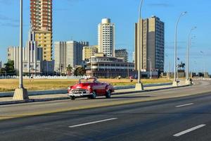 havana, cuba - 15 de janeiro de 2017 - carro clássico dirigindo ao longo do malecon com a casa de las americas ao fundo em havana, cuba. foto