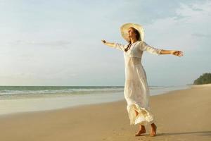 mulher vestindo lindo vestido branco está caminhando na praia durante o pôr do sol foto