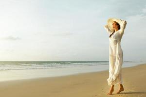 mulher vestindo lindo vestido branco está caminhando na praia durante o pôr do sol foto