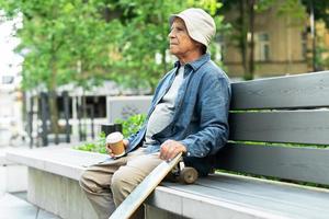 homem idoso com um longboard sentado no banco e tomando café em um parque da cidade foto