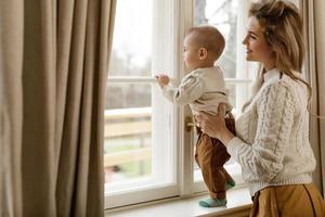 mãe e seu filhinho bebê vestindo suéteres quentes em pé perto da janela foto
