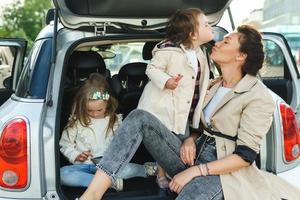 jovem mãe e suas adoráveis filhas sentadas na mala de um carro. foto