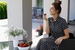 linda mulher vestindo lindo vestido com um padrão de bolinhas sentado em um pátio e comendo morango foto