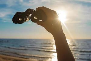 aventura - mão feminina com um binóculo vintage contra a luz do sol brilhante na bela praia com belo céu azul. foto