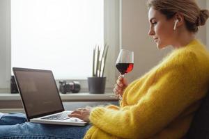 mulher sentada no sofá bebendo vinho tinto enquanto assiste a um filme ou trabalha em seu laptop em casa foto