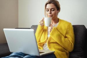 mulher com uma xícara de café quente sentada no sofá e usando o laptop em casa foto