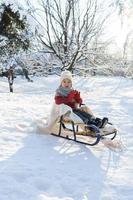 menino da criança sentado no trenó em um parque da cidade de neve durante o dia ensolarado de inverno foto