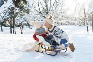 jovem mãe e seu filhinho fofo com trenó retrô em um parque nevado durante o dia ensolarado foto