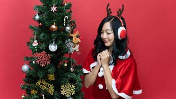 feche o retrato de uma jovem mulher asiática com cabelos compridos vestindo uma fantasia de santo vermelho posando para fazer um desejo sobre fundo vermelho. sorriso de emoção. conceito de ano novo e dia de natal. foto