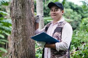 O botânico masculino asiático está inspecionando e registrando as informações do tronco da árvore na área de transferência de papel. conceito, pesquisa, pesquisa de plantas botânicas. conservação florestal e ambiental. foto