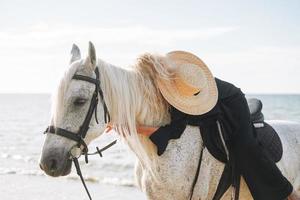 jovem loira em roupas pretas, cavalgando um cavalo branco no fundo da paisagem marinha foto