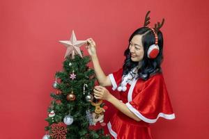 mulher asiática muito sorridente em fantasia de santy e chapéu de papai noel está decorando a árvore de natal em fundo vermelho para o conceito de celebração da temporada foto