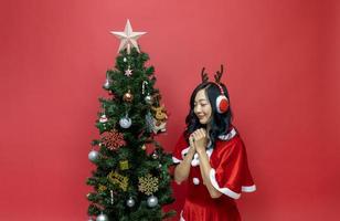 mulher asiática muito sorridente em fantasia de santy e chapéu de papai noel está fazendo um desejo ao lado da árvore de natal em fundo vermelho para o conceito de celebração da temporada foto