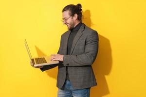 homem barbudo feliz usando óculos está usando computador portátil em fundo amarelo foto