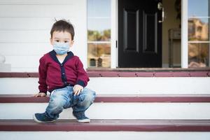 menino melancólico solitário de raça mista na varanda da frente usando máscara facial médica durante a pandemia de coronavírus foto