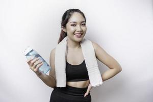 mulher asiática esportiva posando com uma toalha no ombro e segurando uma garrafa de água, sorrindo e relaxando após o treino foto
